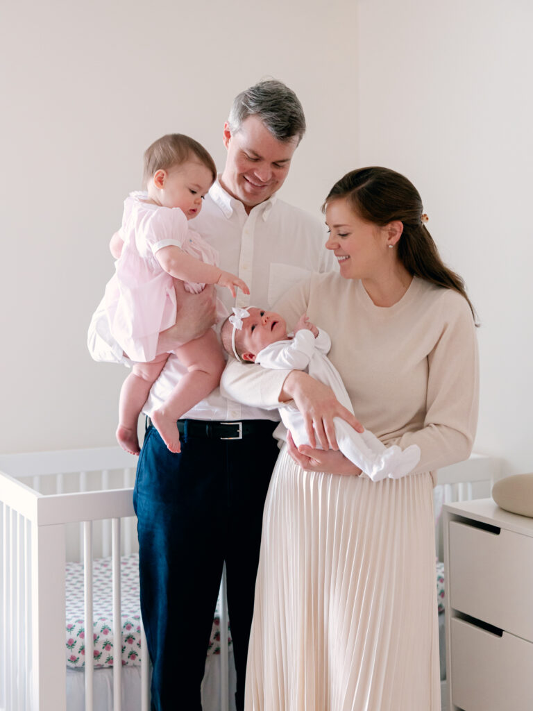 family of 4 with mom bigger sister and dad all looking at newborn baby sister in the nursery